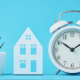 Eine Pflanze, ein Häuschen und ein Wecker stehen vor einem türkisblauem Hintergrund - Zeitpunkt Immobilienverkauf