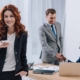 Eine junge Frau steht in einem Büro und ist glücklich über einen neuen Job, im Hintergrund schütteln sich zwei Männer die Hand | Immobilienverkauf bei Jobwechsel