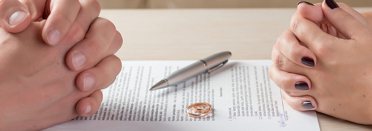 Mann und Frau sitzen sich mit gefaltenten Händen gegenüber an einem Tisch, auf dem Scheidungspapiere, ein Kugelschreiber und ein Ehering liegen | Immobilie Scheidung