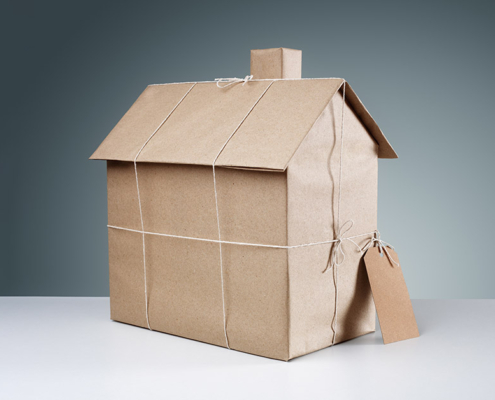 Ein Modell eines Hauses steht in braunem Papier verpackt und mit Schnur zusammengebunden auf einem weißen Tisch vor einem dunklen Hintergrund | Immobilie erben