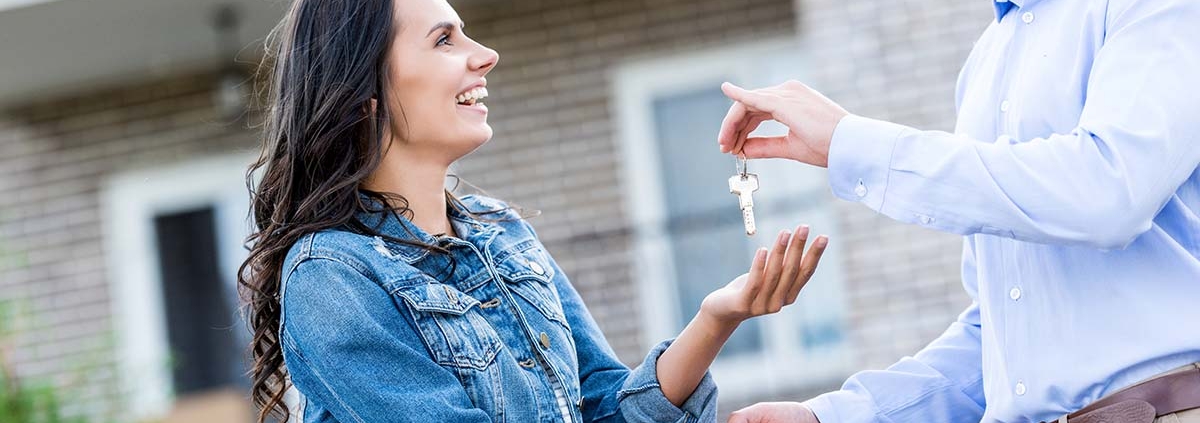 Ein glücklicher Immobilienverkäufer übergibt einer glücklichen Immobilienkäuferin die Hausschlüssel, sie stehen vor dem verkauften Haus | Immobilienkauf