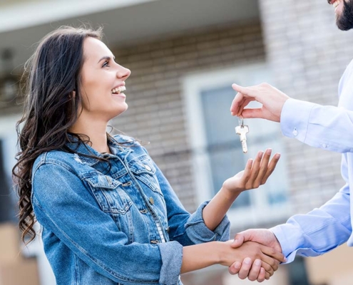 Ein glücklicher Immobilienverkäufer übergibt einer glücklichen Immobilienkäuferin die Hausschlüssel, sie stehen vor dem verkauften Haus | Immobilienkauf